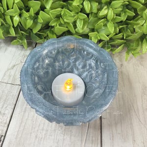 May include: A light blue resin bowl with a floral design, holding a white LED candle with a yellow flame. The bowl is on a light wood surface with green foliage in the background. The text "The Craft Factor" is visible.