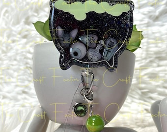 May include: A black cauldron with glitter and various Halloween-themed charms, including an eyeball, a potion bottle, and a sign that says "Do Not Trust". The cauldron is attached to a retractable badge reel with a green and purple beaded chain.