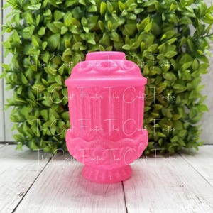 May include: A bright pink decorative lantern with ornate detailing. The lantern has a base, a cylindrical body with vertical lines, and a domed top. The background features green foliage and a wooden surface.