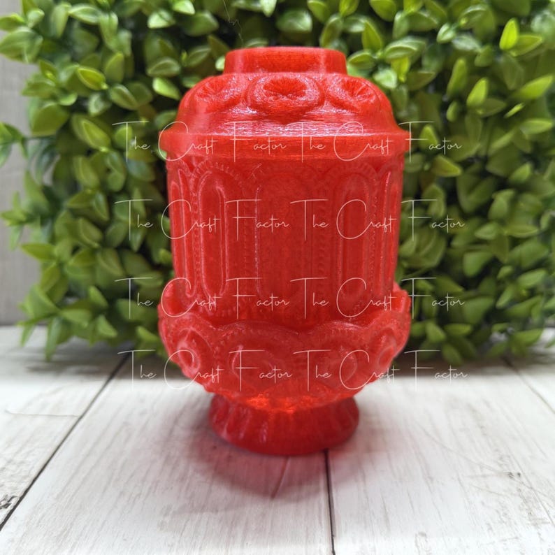 May include: A red decorative jar with a lid. The jar has an ornate design with repeating patterns and a pedestal base. The background features green foliage and a white wooden surface.