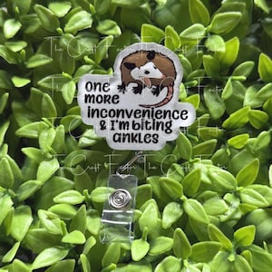 May include: A badge reel featuring a cartoon opossum illustration and the black text one more inconvenience & I'm biting ankles. The badge reel has a clear plastic housing with a retractable cord and a metal clip mechanism for attachment.