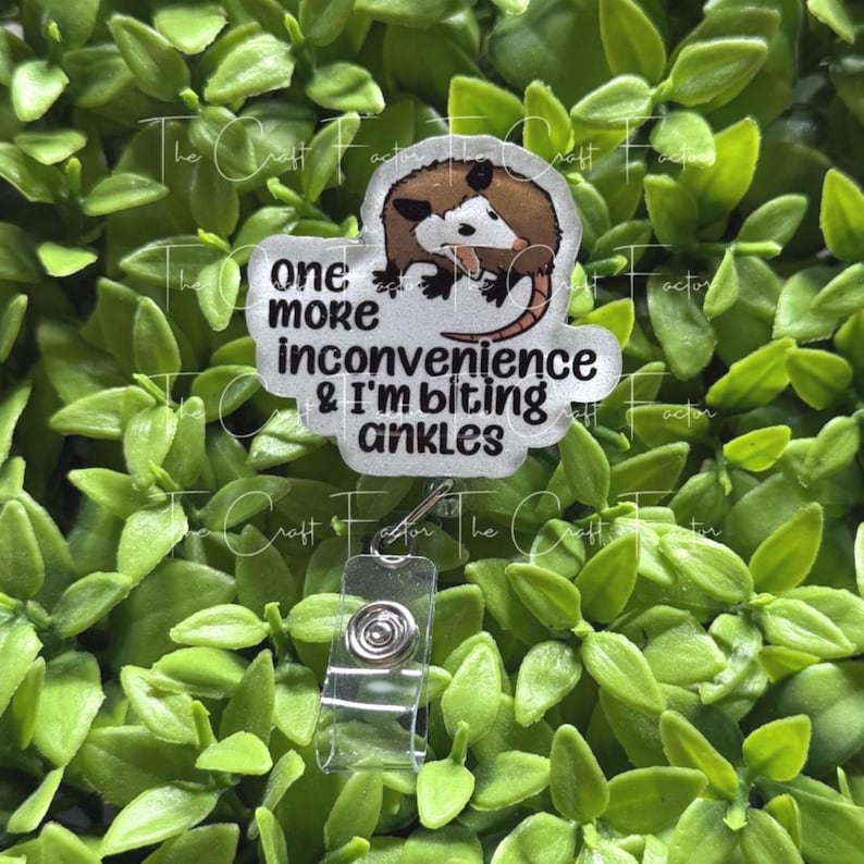 May include: A badge reel featuring a cartoon opossum illustration and the black text one more inconvenience & I'm biting ankles. The badge reel has a clear plastic housing with a retractable cord and a metal clip mechanism for attachment.