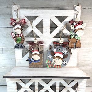 May include: Wooden Christmas ornaments featuring moose designs. Each moose wears a Santa hat and scarf, with some holding items like a mug or snowflake. The ornaments are decorated with red and green ribbons and the text "CHRISTMOOSE".