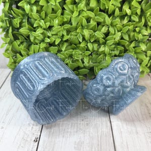 May include: Two blue resin decorative pieces. One is a cup-shaped object with vertical lines, and the other is a detailed, stylized dragon head. The pieces are on a light wood surface with green foliage in the background.