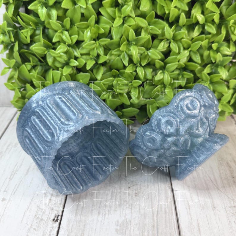 May include: Two blue resin decorative pieces. One is a cup-shaped object with vertical lines, and the other is a detailed, stylized dragon head. The pieces are on a light wood surface with green foliage in the background.