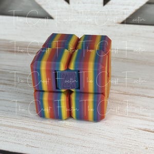May include: A colorful, rainbow-striped fidget cube. The cube is made of multiple connected blocks that can be rotated and manipulated. The cube has a purple center piece and is designed to provide tactile stimulation.