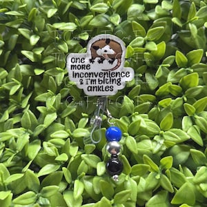 May include: A badge reel featuring a cartoon opossum illustration and the text "one more inconvenience & i'm biting ankles". The badge reel includes a clear plastic strap and a vertical dangle of three beads in blue, silver, and black colours.