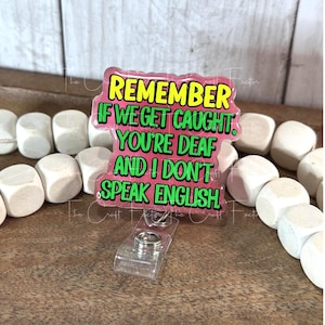 May include: A pink and yellow badge reel with the text "REMEMBER IF WE GET CAUGHT. YOU'RE DEAF AND I DON'T SPEAK ENGLISH" in green and white. The badge reel has a clear plastic clip and a silver metal reel.