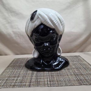 May include: A glossy ceramic head vase featuring a person with black skin wearing a white turban with a dark accent piece at the center. The figure has defined facial features and large white teardrop-shaped earrings against a black base.