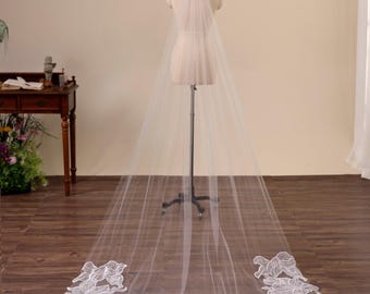 May include: A long, sheer white bridal veil with a floral lace border. The veil is displayed on a mannequin, showcasing its length and delicate design. The lace detailing is concentrated at the bottom edge, adding a touch of elegance.