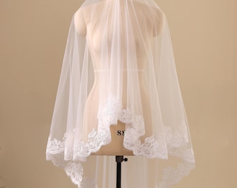 May include: A white wedding veil made of sheer tulle fabric, featuring an intricate floral lace trim along the scalloped edges. The veil is draped over a mannequin, showcasing its layered design and delicate embroidery that frames the lower section.