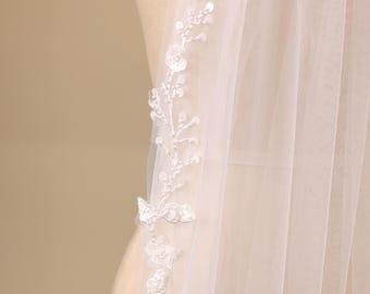 May include: A sheer white tulle veil featuring an intricate embroidered vine pattern along the edge. The design includes delicate white floral motifs and small shimmering sequins that catch the light, creating a subtle texture on the lightweight fabric.