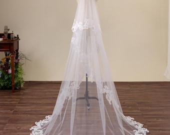 May include: A long, two-tier white bridal veil made of sheer tulle fabric. The veil features intricate white floral lace embroidery along the edges of both tiers and a scalloped lace trim that extends around the entire circular hemline.