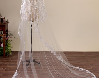 May include: A long, floor-length white bridal veil draped over a mannequin. The veil features intricate white floral lace embroidery with shimmering beadwork and sequins throughout the top section and along the scalloped, rounded bottom edge.