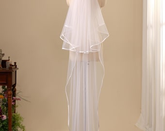 May include: A two-tier white wedding veil featuring a delicate satin ribbon trim along the edges. The sheer tulle fabric is gathered at the top and drapes down in a long, flowing silhouette, secured to a mannequin for a clear display of its length.