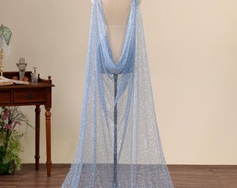 May include: A floor-length, light blue cape made of sheer, sparkling fabric. The garment features a deep, open back design with wide shoulder straps that drape down to the floor, creating a flowing silhouette on a dress form mannequin.