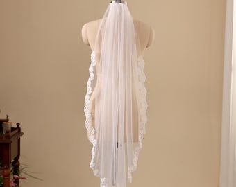 May include: A white bridal veil made of sheer tulle fabric, featuring a scalloped lace trim with floral embroidery along the edges. The veil is draped over a mannequin, showcasing a layered design that falls vertically in a soft, flowing manner.