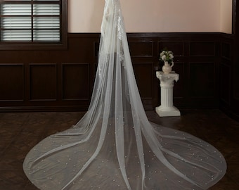 May include: A long white tulle wedding veil with pearl embellishments. The veil is draped over a mannequin and has a scalloped edge.