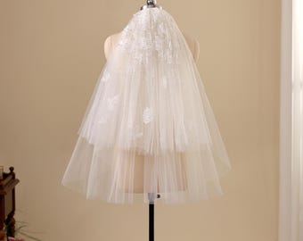May include: A two-tier white tulle wedding veil featuring delicate floral lace appliques and scattered small beads. The veil is draped over a mannequin, showcasing a layered design with a gathered top and sheer fabric flowing downwards.