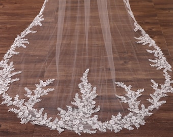 May include: A sheer white wedding veil with a delicate floral lace trim along the edge. The lace features detailed floral and leaf patterns. The veil is spread out on a wooden surface.