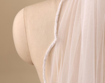 May include: A sheer white bridal veil featuring a delicate edge trim composed of small, tightly clustered white beads. The lightweight tulle fabric drapes vertically, showcasing the intricate beaded detailing along the finished border of the veil.