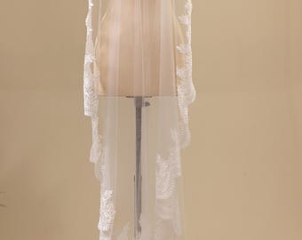 May include: A long, sheer white bridal veil draped over a mannequin. The veil features intricate white floral lace embroidery along the edges, with a scalloped hemline that extends down the length of the fabric, creating a delicate, layered appearance.