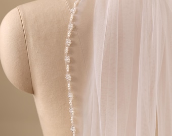 May include: A sheer white bridal veil featuring a delicate edge trim composed of small round white pearls and clear faceted beads arranged in a repeating floral pattern along the entire length of the tulle fabric draped over a mannequin.