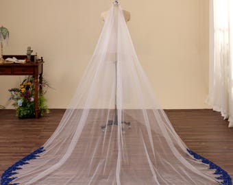 May include: A long, white bridal veil with a deep blue lace trim. The veil is displayed on a mannequin, showcasing its length and delicate design. The blue lace trim adds a pop of color and intricate detail to the classic white veil.