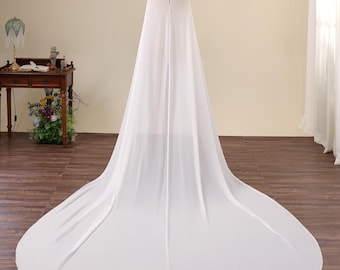 May include: A floor-length white cape made of a sheer, lightweight fabric. The garment features a rounded neckline with a small button closure at the top and flows down into a wide, circular train that spreads across the floor behind the mannequin.