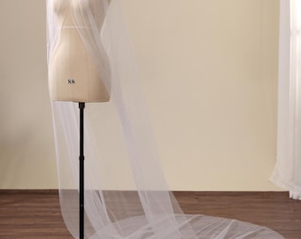 May include: A floor-length white tulle cape featuring a high neckline adorned with silver-toned embellishments. The sheer, lightweight fabric drapes from the shoulders, extending into a long train that pools on the floor, displayed on a mannequin.