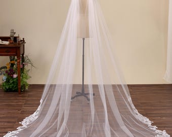 May include: A floor-length white bridal cape made of sheer tulle fabric. The cape features a wide, scalloped hemline decorated with intricate white floral embroidery and lace detailing that extends along the entire bottom edge of the garment.