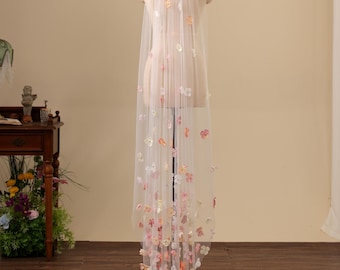May include: A long, sheer white bridal veil draped over a mannequin, featuring scattered three-dimensional fabric flower appliqués in shades of pink, peach, yellow, and white that increase in density toward the bottom hem of the veil.
