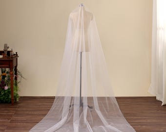 May include: A long, floor-length wedding veil made of sheer, translucent white tulle fabric. The veil is draped over a mannequin, extending from the top down to the floor, creating a wide, circular train that spreads out across the ground.