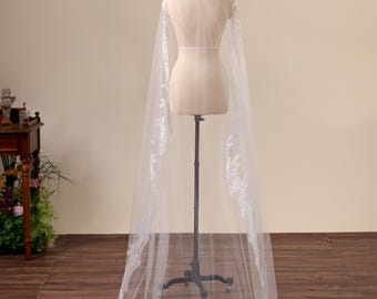 May include: A long, floor-length white wedding veil draped over a mannequin. The veil is made of sheer tulle fabric and features intricate white floral lace embroidery along the edges and a decorative lace applique at the top.