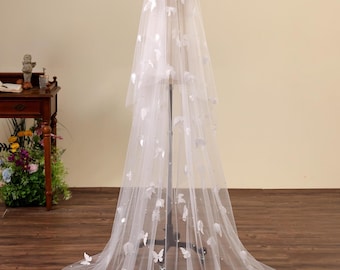 May include: A long, sheer white wedding veil featuring scattered three-dimensional white butterfly appliqués and small, evenly spaced white pearls throughout the fabric. The veil is displayed on a mannequin, cascading down to the floor in a circular train.