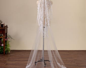 May include: A long white wedding veil draped over a mannequin, featuring intricate white floral lace embroidery at the top and along the scalloped hemline. The sheer tulle fabric extends to the floor, creating a flowing, elegant silhouette.