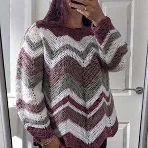 Puede incluir: Un suéter de crochet hecho a mano con un patrón de chevrón en tonos blanco, gris y burdeos. El suéter tiene mangas largas y un cuello redondo. El estilo es casual y cómodo.