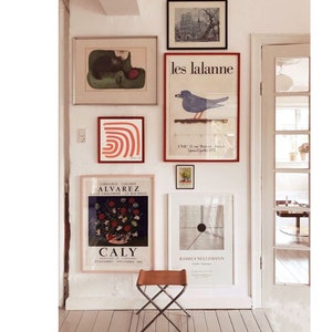 May include: A white wall with a collection of framed artwork, including a black and white illustration of a cat, a red and white abstract design, a poster with a bird illustration and the text "les lalanine", and a poster with the text "CALY" and a floral illustration.