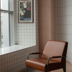 May include: A brown leather armchair with a metal frame and wooden armrests sits in a room with white tiled walls and a window. A framed print of a Japanese flower arrangement hangs on the wall.