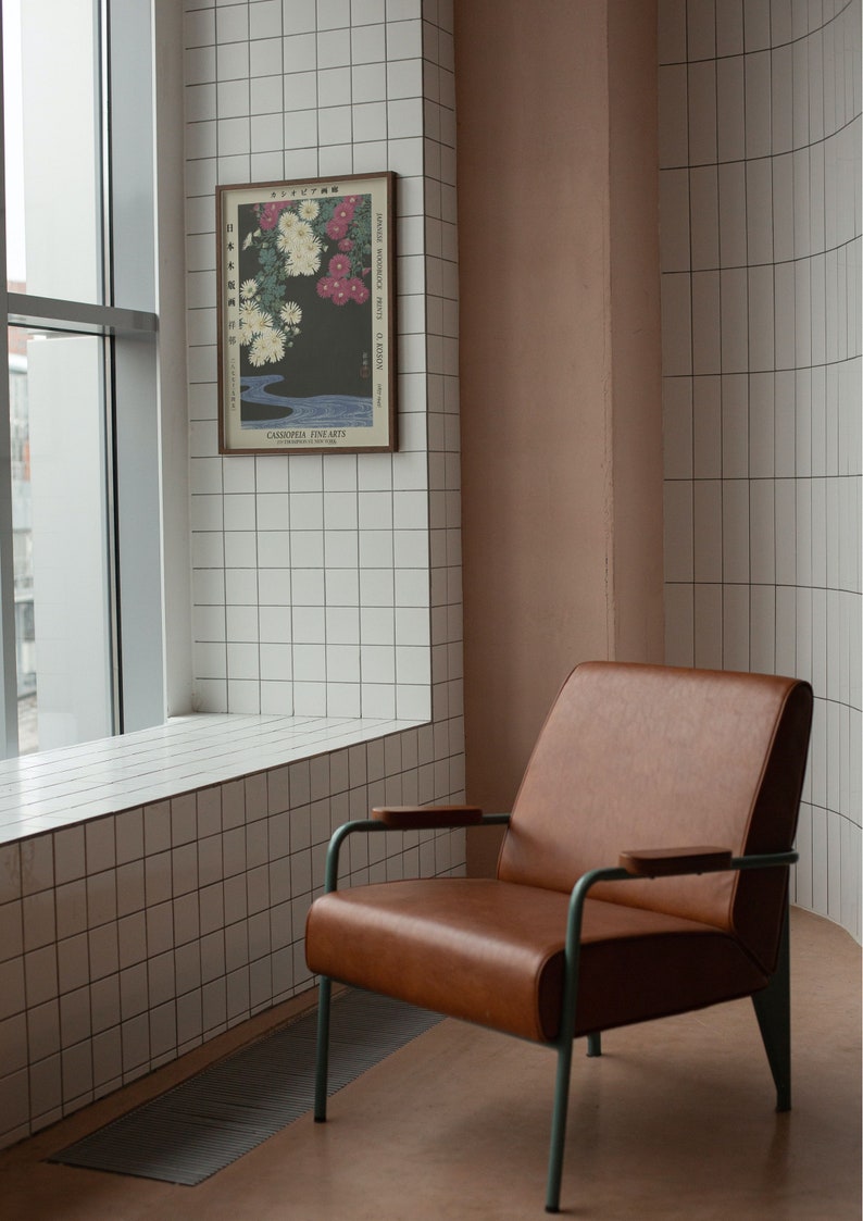 May include: A brown leather armchair with a metal frame and wooden armrests sits in a room with white tiled walls and a window. A framed print of a Japanese flower arrangement hangs on the wall.