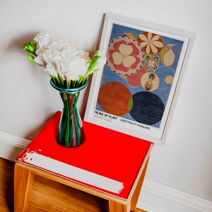 May include: A red side table with a white painted stripe on the top. A white framed poster with a blue background and colourful abstract circles hangs on the wall above the table. The poster reads "Hilma af Klint - Abstract Pionjar" and "Moderna Museet". A vase with white flowers sits on the table.