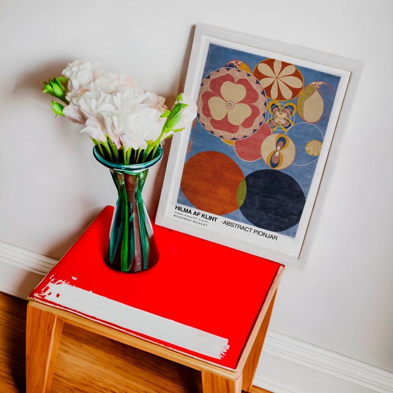 May include: A red side table with a white painted stripe on the top. A white framed poster with a blue background and colourful abstract circles hangs on the wall above the table. The poster reads "Hilma af Klint - Abstract Pionjar" and "Moderna Museet". A vase with white flowers sits on the table.