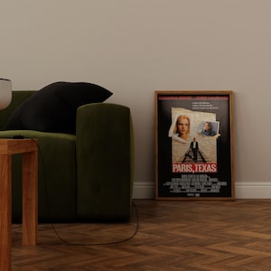 May include: A framed movie poster for "Paris, Texas" with a black background and images of the actors. The poster is in a wooden frame and is leaning against a white wall. A green sofa and wooden table are in the foreground.