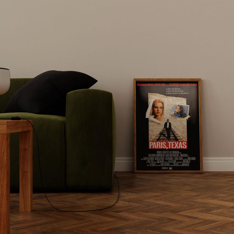 May include: A framed movie poster for "Paris, Texas" with a black background and images of the actors. The poster is in a wooden frame and is leaning against a white wall. A green sofa and wooden table are in the foreground.