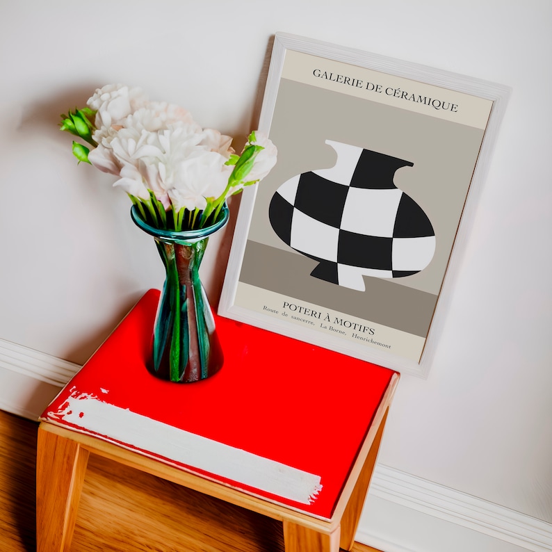 May include: A white framed poster with black and white checkered pottery on a beige background. The poster reads "Galerie de C&eacute;ramique" and "Poterie &agrave; Motifs". The poster is sitting on a red table with a vase of white flowers.
