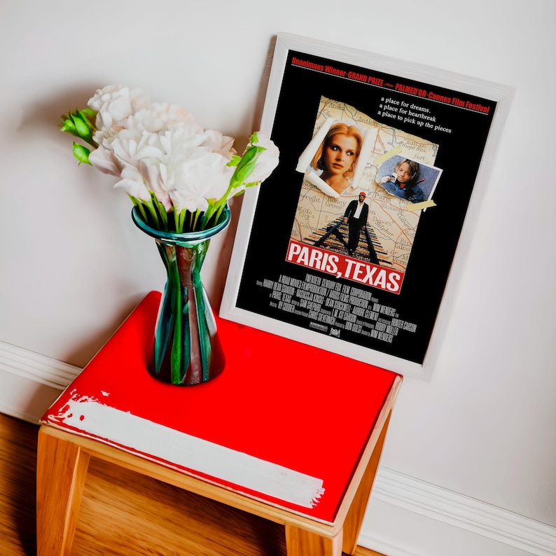 May include: A framed movie poster for "Paris, Texas" with a white border rests on a small red table. A glass vase with green and brown accents holds white flowers. The table has wooden legs and a white stripe.