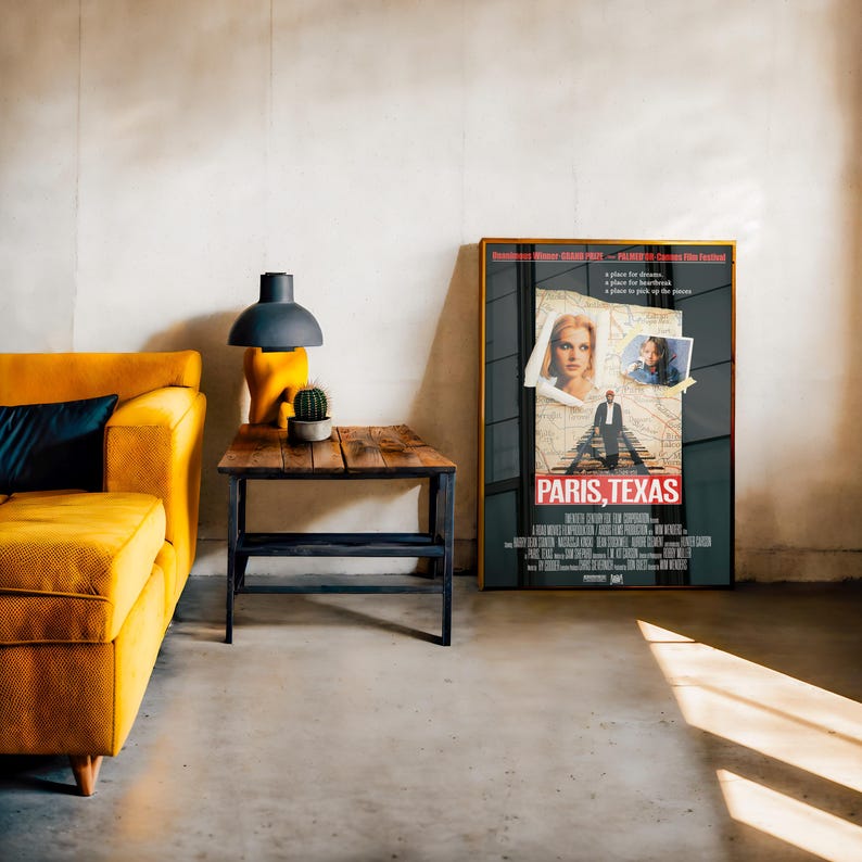 May include: A living room scene featuring a mustard yellow sofa, a wooden side table with a black metal frame, and a framed movie poster. The poster is for the film "Paris, Texas". A black lamp and a small cactus are on the side table.