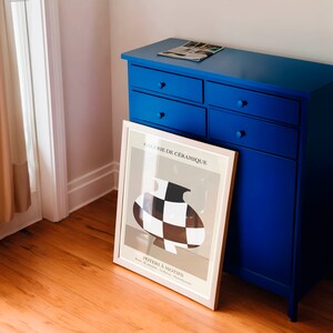 May include: A blue dresser with four drawers and a white framed poster with a black and white checkered design. The poster reads "Galerie de Ceramique" and "Poterie a Motifs".