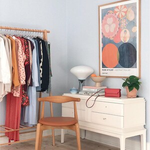 May include: A white dresser with three drawers and a wooden top. A brown leather chair sits in front of the dresser. A white lamp with a white shade sits on the dresser. A framed poster with the text "HILMA AF KLINT - ABSTRAKT PUNKTER" hangs on the wall. A potted plant sits on the dresser.
