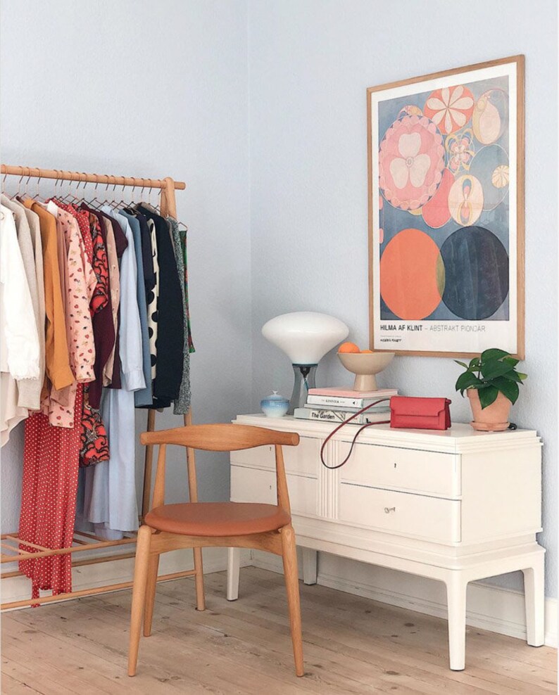 May include: A white dresser with three drawers and a wooden top. A brown leather chair sits in front of the dresser. A white lamp with a white shade sits on the dresser. A framed poster with the text "HILMA AF KLINT - ABSTRAKT PUNKTER" hangs on the wall. A potted plant sits on the dresser.